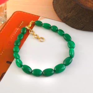 Puede incluir: Una pulsera de cuentas verdes con cuentas de forma ovalada. La pulsera tiene un cierre y una cadena extensora de color dorado. Las cuentas son de un color verde vibrante y sólido. La pulsera se muestra sobre una superficie blanca.