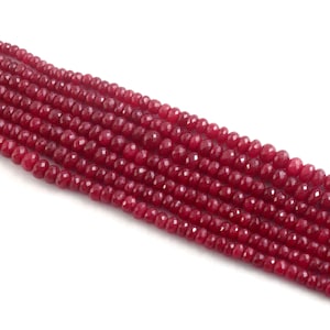 May include: Strands of faceted, deep red beads. The beads are small and uniform in size, with a polished, reflective surface. The beads are strung together, creating a flowing, textured appearance. The background is white.