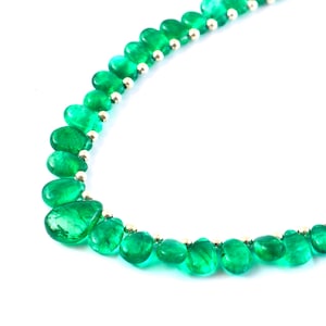 Puede incluir: Un collar con cuentas verdes vibrantes en forma de lágrima, intercaladas con pequeñas cuentas doradas. El collar se presenta sobre un fondo blanco limpio, resaltando el color y la forma de las piedras preciosas.