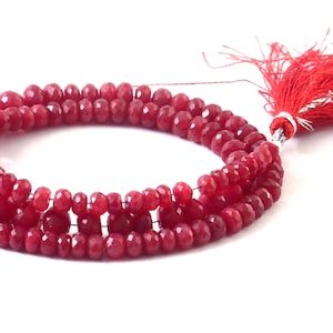 Puede incluir: Hebras de cuentas facetadas de piedras preciosas de color rojo intenso. Las cuentas están ensartadas, formando múltiples filas. Una borla roja con detalles plateados está unida al final de las hebras. El fondo es blanco.