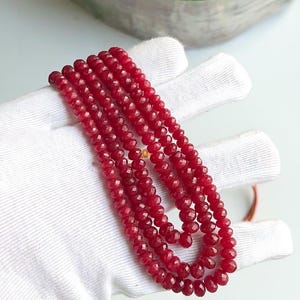 以下が含まれることがあります： 深紅色のファセット宝石のネックレスのコレクション。ビーズはつながっており、複数のストランドを形成しています。ネックレスは白い表面に表示され、色とデザインが強調されています。宝石は均一なサイズと形状のようです。