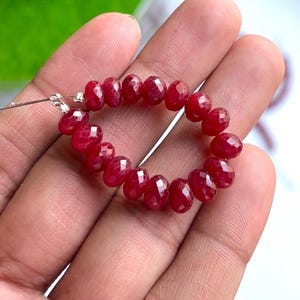 Può includere: Un filo di perline di pietre preziose rosse scuro sfaccettate. Le perline sono infilate su un filo sottile color argento, formando una forma circolare. Le pietre preziose hanno una superficie lucida e riflettente e sono tenute in una mano.