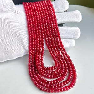 May include: A collection of six strands of faceted, deep red gemstone beads. The beads are small and uniform in size, creating a rich, textured appearance. The strands are draped over a white surface, showcasing the color and shine of the gemstones.