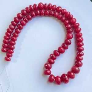 Puede incluir: Dos hilos de cuentas de piedras preciosas facetadas de color rojo intenso. Las cuentas de forma ovalada están ensartadas en un fino cordón transparente. Las cuentas tienen un aspecto brillante y pulido, y están dispuestas en un patrón curvo sobre un fondo blanco.