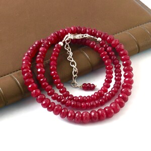 May include: A layered necklace featuring faceted, deep red gemstones. The necklace has a silver-colored clasp and extender chain. The gemstones are strung together to create a multi-strand design. The necklace is displayed on a brown surface.