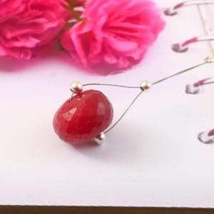 Peut inclure: Un collier avec un pendentif en pierre précieuse rouge profond et des accents argentés. Le pendentif est de forme arrondie et facettée. En arrière-plan, des fleurs roses et une surface blanche.