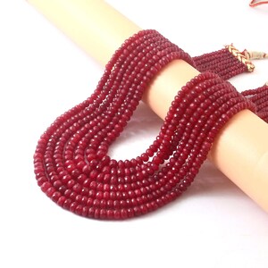 Puede incluir: Un collar de varias hebras con cuentas facetadas de color rojo intenso. El collar está drapeado sobre una forma cilíndrica de color claro, mostrando el color y la textura de las cuentas. El collar tiene una borla de cuentas a juego.
