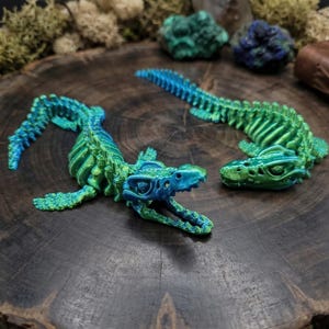 Op de afbeelding: Twee gearticuleerde, 3D-geprinte drakenfiguren in blauwe en groene tinten liggen op een donker houten oppervlak. De draken hebben gedetailleerde skeletstructuren en zijn ontspannen gepositioneerd. Decoratief mos en stenen zijn op de achtergrond.