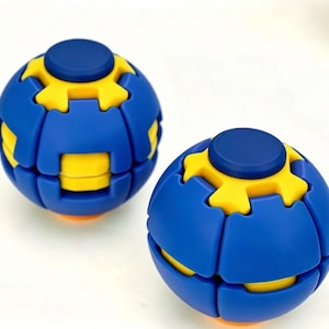May include: Two spherical fidget puzzles featuring a blue and yellow interlocking gear design. Each sphere has a smooth blue surface with yellow accents and a central blue circular button on top, creating a mechanical look with segmented parts.