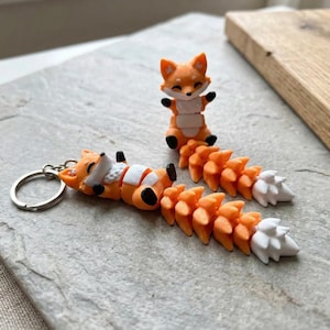 May include: Orange fox keychain and figurine. The keychain features a fox lying down with a white belly and tail. The figurine is a standing fox with its arms raised. Both have orange and white tails.