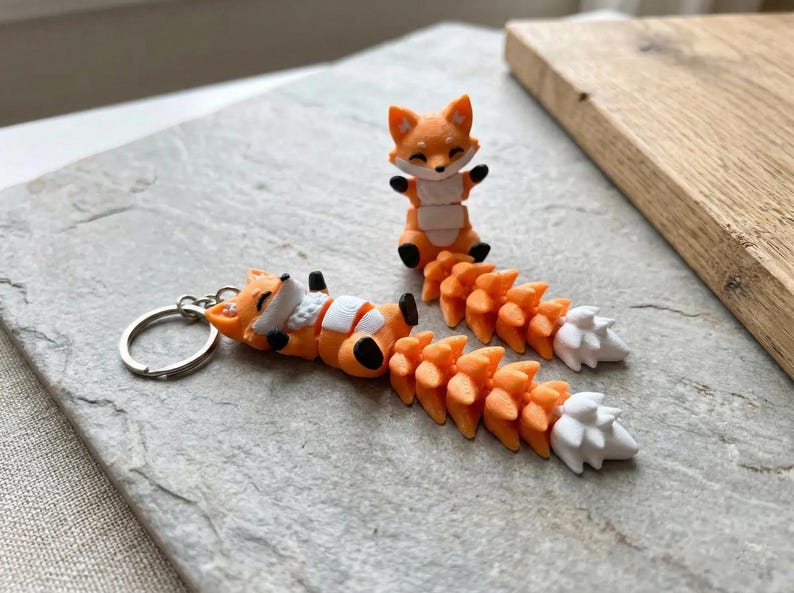 May include: Orange fox keychain and figurine. The keychain features a fox lying down with a white belly and tail. The figurine is a standing fox with its arms raised. Both have orange and white tails.