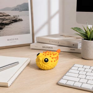May include: A yellow pufferfish-shaped toy with orange spots and a small orange tail. The toy has large black eyes and a textured surface. It is sitting on a wooden desk near a keyboard and a potted plant.