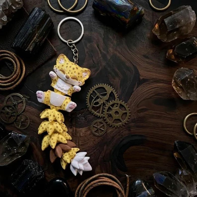May include: A yellow and white keychain featuring a cartoon leopard with closed eyes, suspended from a silver keyring. The leopard has black spots and is attached to a decorative tail. The background includes gears, crystals, and leather straps.