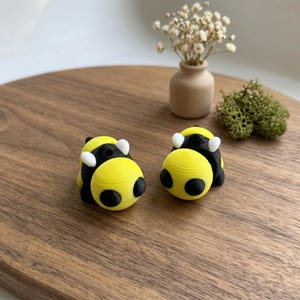Puede incluir: Dos pequeñas figuras de abeja amarillas y negras con detalles blancos. Las figuras de abeja tienen forma redondeada con ojos negros y se exhiben sobre una superficie de madera. Un pequeño jarrón con flores secas y un trozo de musgo verde están en el fondo.