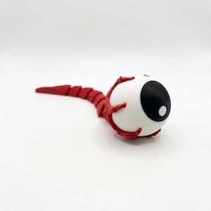 May include: A 3D-printed, stylized eyeball sculpture. The eye features a white sclera, a black iris, and a white pupil. Red, vein-like structures extend from the eye, connecting to a segmented, red tail. The sculpture is set against a white background.