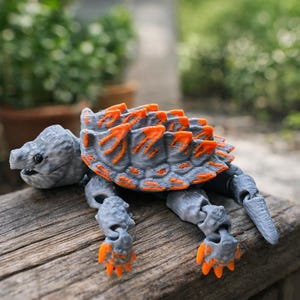May include: A gray and orange articulated turtle toy. The turtle has a gray body with orange accents on its shell and feet. The toy is made of plastic and is posed on a wooden surface.