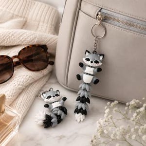 May include: Two raccoon keychains, one attached to a silver keyring, the other is separate. The raccoons are gray, black, and white, with a detailed tail. A pair of tortoiseshell sunglasses and a light gray purse are in the background.