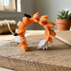 May include: A 3D-printed orange fox toy with black and white accents, posed in an arch. The fox is on a wooden surface, with a small potted succulent and a roll of twine in the background. The fox's tail is white.