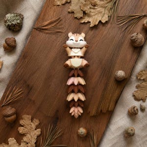 May include: A whimsical figurine of a chipmunk with outstretched arms, atop a stack of stylized leaves in shades of pink and brown. The chipmunk is tan with brown stripes. Acorns and dried leaves surround the figurine on a wooden surface.