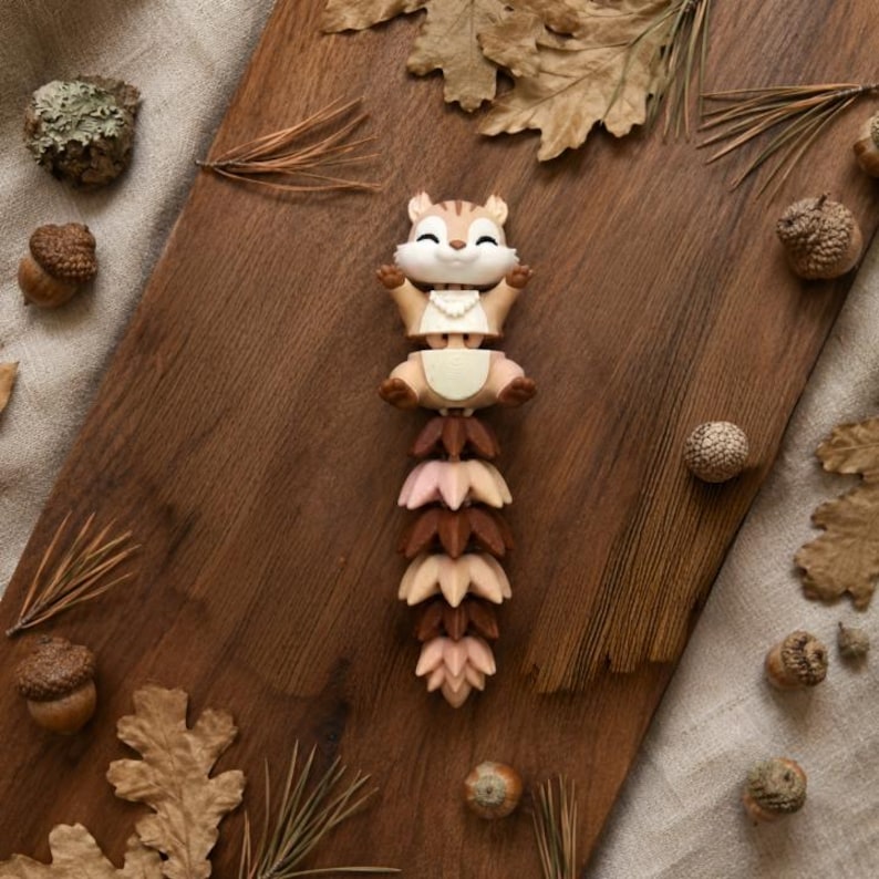 May include: A whimsical figurine of a chipmunk with outstretched arms, atop a stack of stylized leaves in shades of pink and brown. The chipmunk is tan with brown stripes. Acorns and dried leaves surround the figurine on a wooden surface.