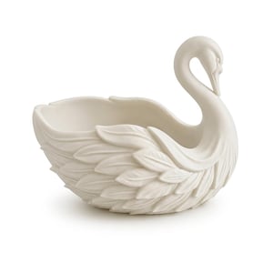 Puede incluir: Un cuenco decorativo de cerámica blanca con forma de cisne. El cisne tiene un cuello largo y curvado y texturas detalladas en forma de plumas en su cuerpo. El cuenco está diseñado para guardar objetos pequeños.