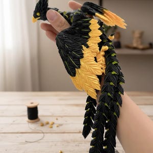 May include: A detailed bird sculpture with black and yellow feathers. The bird has a yellow beak and is held in a hand. The sculpture is on a wooden surface with sewing supplies in the background. The bird's tail feathers are long and detailed.