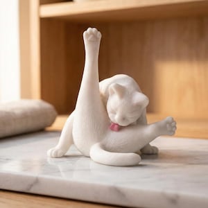 Może przedstawiać: Biała ceramiczna figurka kota w zabawnej pozie, czyszczącego się. Kot jest przedstawiony z podniesioną łapą i wystawionym językiem. Figurka stoi na marmurowej powierzchni.