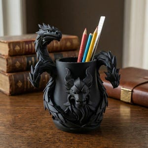Könnte beinhalten: Ein schwarzer Stifthalter im Drachen-Design mit drei Drachenköpfen und einem detaillierten, strukturierten Design. Der Halter enthält mehrere farbige Bleistifte und ein Lineal. Der Stifthalter besteht aus einem dunklen Material und steht auf einer Holzoberfläche.