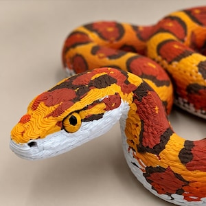 May include: A 3D printed snake figurine featuring a textured surface with a vibrant pattern of orange, red, and dark brown blotches. The snake has a white underbelly and a detailed head with black eyes, showcasing visible layer lines from the printing process.