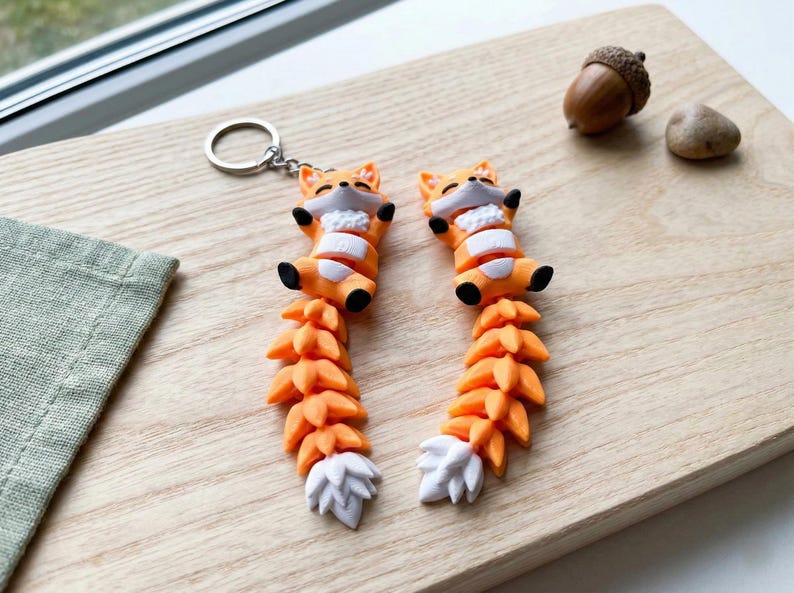 May include: Two orange fox keychains with white and black accents, lying on a wooden surface. The keychains have a detailed tail design and a silver keyring. An acorn and a small stone are also on the wooden surface.