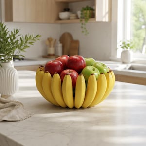 Op de afbeelding: Een decoratieve fruitschaal in de vorm van een tros gele bananen, gevuld met een mix van verse rode en groene appels. De schaal heeft een gebogen, gesegmenteerd ontwerp dat het uiterlijk nabootst van individuele bananen in een cirkel.
