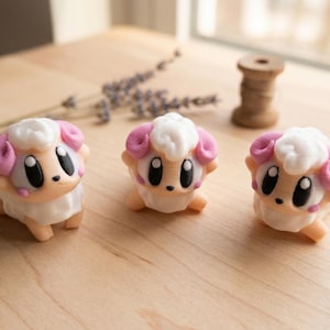 Könnte beinhalten: Drei kleine Schaf-Figuren im Cartoon-Stil. Jede hat einen weißen, flauschigen Kopf, rosa Hörner und große schwarze Augen. Die Schafe sind hellbraun mit weißen Bäuchen und stehen auf einer hellen Holzoberfläche.