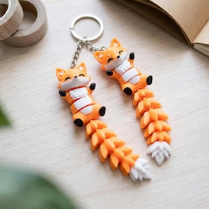 May include: Two orange fox keychains with white and black accents. The foxes have outstretched arms and long, detailed tails. The keychains are on a silver chain and ring. A cute and whimsical accessory.