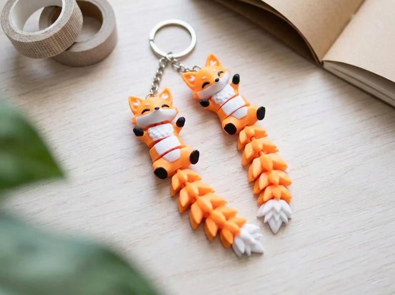 May include: Two orange fox keychains with white and black accents. The foxes have outstretched arms and long, detailed tails. The keychains are on a silver chain and ring. A cute and whimsical accessory.