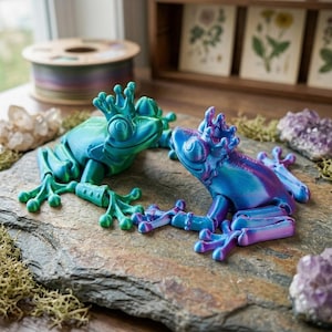 Op de afbeelding: Twee decoratieve kikkerbeeldjes in blauwe en groene tinten, elk met een kroon. De kikkers staan op een stenen oppervlak, met mos en kristallen op de achtergrond. De kikker links is groen en blauw, en de kikker rechts is blauw en paars.