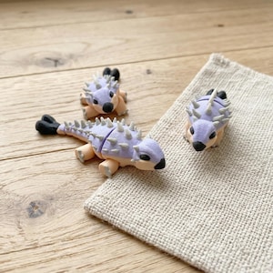 May include: Three small, handcrafted dinosaur figurines. The dinosaurs are light purple with tan bellies and black accents. They have spiky backs and are arranged on a wooden surface and a beige linen napkin.