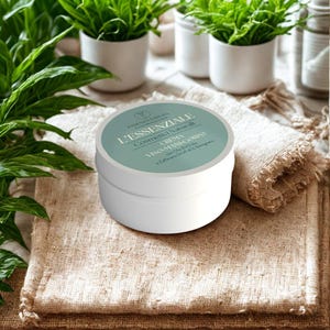 Può includere: Un barattolo cosmetico bianco con coperchio verde chiaro, con testo in italiano. Il barattolo è etichettato "L'ESSENZIALE" e "Cosmetici Naturali". Il barattolo è su un tessuto strutturato, beige, con piccole piante in vaso sullo sfondo.