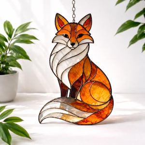 Op de afbeelding: Gebrandschilderd glazen zonnevanger van een zittende vos. De vos heeft tinten oranje, wit en bruin, met een zwarte omtrek. Hij hangt aan een ketting. Het ontwerp van de vos is geometrisch, met donkere lijnen.