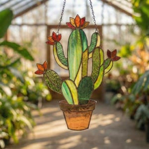Op de afbeelding: Gebrandschilderde cactus zonnevanger met een bruine pot. De cactus heeft groene, gele en oranje tinten, met een ketting om op te hangen. Het ontwerp omvat een centrale stengel en zijarmen, met oranje bloemaccenten.