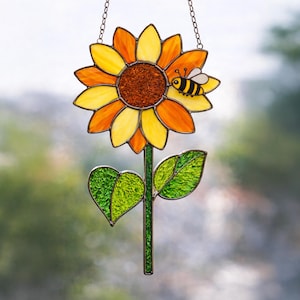 May include: Stained glass sunflower with yellow, orange, and brown petals, a green stem, and two green leaves. A small bee with black and yellow stripes is perched on one of the petals. The suncatcher hangs from a chain.