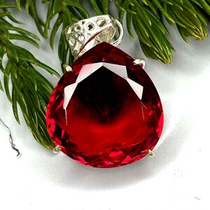 May include: A teardrop-shaped, faceted red gemstone pendant with a silver-colored bail. The pendant is set against a backdrop of green foliage. The gemstone has a deep, rich color and a brilliant cut, reflecting light.