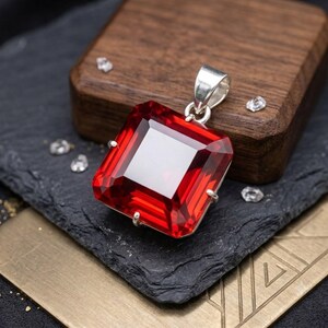 May include: A square-cut, deep red gemstone pendant set in a silver-toned metal prong setting. The pendant features a faceted surface that reflects light, with a silver-toned bail at the top for attachment to a chain or cord necklace.