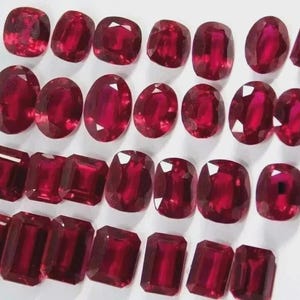 May include: A selection of deep red gemstones in diverse forms, such as oval, cushion, and emerald cuts. The gems are presented on a white surface, highlighting their intense colour and transparency. These gemstones are suitable for jewellery making.
