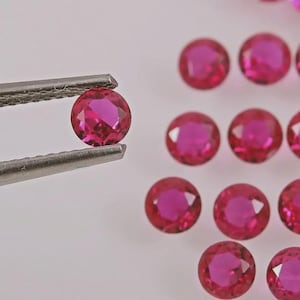 May include: A collection of small, round, faceted, vibrant pink gemstones. One individual gemstone is held securely by a pair of metal tweezers, showcasing its circular cut and reflective surface, while several other identical stones are scattered nearby.