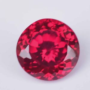 May include: A round, faceted gemstone with a vibrant, deep red color. The stone features a brilliant cut with numerous triangular and kite-shaped facets that reflect light, creating a complex pattern of light and dark red tones across its surface.