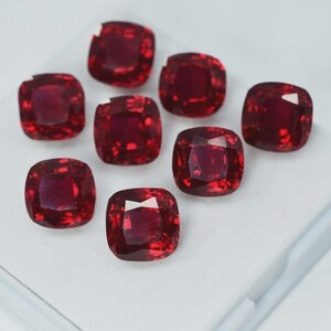 Peut inclure: Huit pierres précieuses de couleur rouge profond, taillées en coussin, sont disposées sur une surface blanche. Les gemmes ont une forme carrée aux coins arrondis et présentent une taille brillante à facettes, reflétant la lumière.
