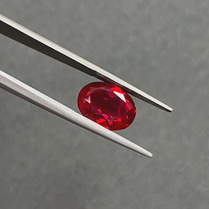 May include: A faceted, oval-shaped, deep red gemstone is held by silver tweezers against a gray background. The gem's polished surfaces reflect light, showcasing its brilliance. The tweezers provide a close-up view of the gemstone.