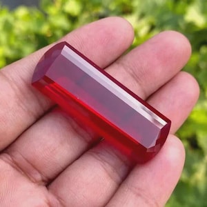 以下が含まれることがあります： ファセット加工された長方形の深紅色の宝石。 宝石は手で持たれており、背景はぼやけた緑色です。 宝石は研磨された反射面を持ち、ファセットは光と影の遊びを生み出します。 全体的な形状は円筒形です。