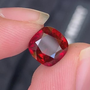 Könnte beinhalten: Ein Edelstein im Kissenschliff mit einer tiefen, leuchtend roten Farbe und einer facettierten Oberfläche, die das Licht reflektiert. Der Stein hat eine quadratische Form mit abgerundeten Ecken und eine klare, polierte Oberfläche.
