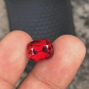 Puede incluir: Una vibrante gema roja, tallada en cojín, sostenida entre los dedos. Las facetas de la gema reflejan la luz, mostrando su claridad y rico color. La piedra parece ser un rubí o una piedra preciosa similar.